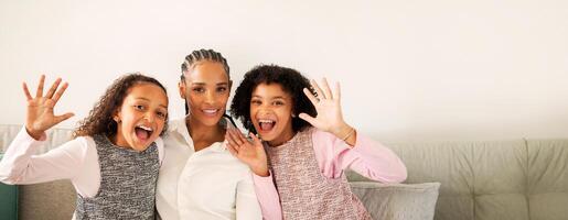A black mother sits on the couch with her two daughters, all smiling and playfully waving their hands. They're enjoying a moment of happiness together in a cozy living room setting. photo