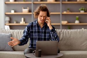 Discontented Man Talking On Phone Using Laptop Having Problem With Computer Or Internet Connection Sitting At Home. Displeased Customer Calling To Hotline Service. Selective Focus photo