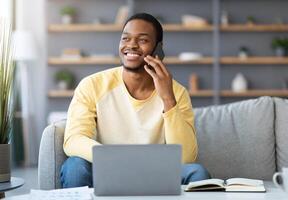 Joyful black guy freelancer having phone conversation while working from home, using laptop, smiling and looking at copy space. Young african american businessman talking with clients on mobile phone photo