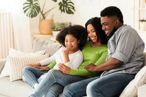 Happy black father, mother and their little daughter relaxing with smartphone at home together, sitting on couch, watching videos or making call photo