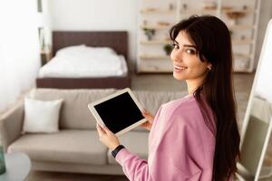 Portrait of smiling young woman holding digital tablet with black empty screen for mockup template, looking back at camera, standing in modern living room. Technology, Automation, Smart Home Concept photo