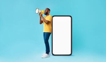 Happy young black guy shouting into megaphone, standing near big smartphone with empty screen, promoting your mobile app or website, offering mockup space for advertisement, blue background photo