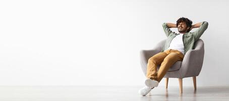 Relaxed curly bearded dark-skinned guy in casual outfit sitting at armchair with arms behind head, looking at copy space for advertisement or text and smiling over white studio background, panorama photo