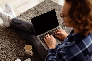 Back-View Of Guy Using Laptop With Blank Screen For Website Advertisement Sitting On Floor At Home. Man Browsing Internet On Computer. Online Technology, Distant Work Concept. Mockup, High Angle Shot photo