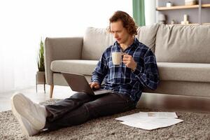 Freelance Work. Serious Guy Using Laptop Computer Working Online And Drinking Coffee Sitting On Floor At Home. Male Freelancer Browsing Internet Indoors. Internet And Technology, Remote Job photo