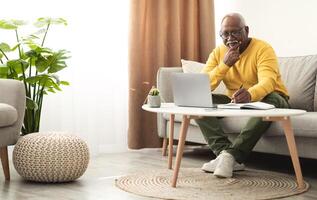 Senior Black Male Using Laptop Working Online And Taking Notes Sitting On Sofa At Home. Mature African American Man Browsing Internet On Computer. Freelance Career And Lifestyle photo
