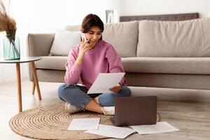 Business, Technology And Communication Concept. Young smiling woman making phone call while looking at document with graphs, busy employee sitting on floor at home office with laptop computer photo