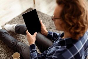 Hipster Guy Using Digital Tablet Computer With Empty Screen Browsing Internet Sitting On Floor At Home. Website Advertisement Concept. Selective Focus, Rear View, Mockup photo