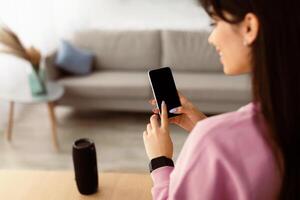 Over The Shoulder View Of Smiling Unrecognizable Young Woman Holding And Using Cell Phone, Touching Empty Mockup Screen With Finger, Standing In Living Room At Home. Selective Focus, Template photo