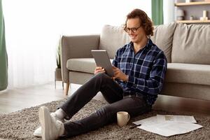 Cheerful Man Using Tablet Computer Browsing Internet Sitting On Floor At Home. Guy Watching Movie Indoor. Application Or Website Advertisement, Gadget And Technology Concept. Side View photo