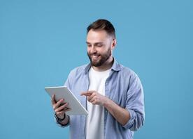 Joyful bearded man using tablet computer for online work or studies on blue studio background. Positive teacher giving remote lesson. Blogger creating new post for social network photo