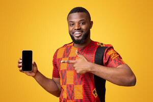Positive african american man pointing at empty smartphone screen, recommending new mobile application, yellow studio background photo