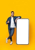 Happy arab guy leaning on huge smartphone with empty screen and showing thumb up, mockup, yellow studio background, full length shot, copy space, mobile application, advertisement concept photo