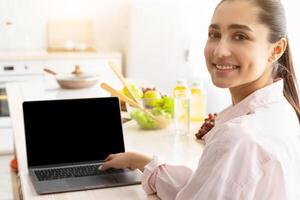 Portrait Of Cheerful Young Woman Using Laptop With Black Empty Screen Sitting At Table In Modern Kitchen At Home. Smiling Woman Looking Back At Camera, Browsing Website With Healthy Recipes, Mockup photo