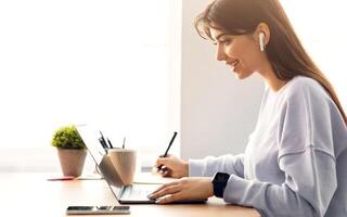 Distance Education. Profile of smiling woman in wireless headphones sitting at desk, using laptop and writing in notebook, taking notes, watching tutorial, lecture or webinar, studying online at home photo