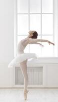 Classical Ballet dancer side view. Beautiful graceful ballerine practice ballet positions in tutu skirt near large window in white light hall. Ballet class training, high-key soft toning. photo