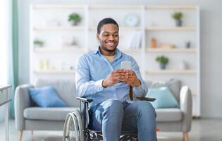 Disabled black man in wheelchair using smartphone, browsing web or watching movie at home. Handicapped young guy checking social media, speaking to friend on mobile device photo