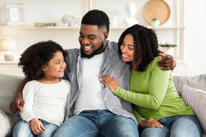 Loving Black Man Embracing His Wife And Daughter While Sitting On Couch At Home, Happy Family Bonding Together photo
