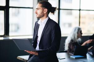 A man in a suit stands with a laptop in hand, engaged in a meeting within a bright, contemporary office space. His colleagues are interacting in the background. photo