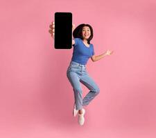 Happy asian young woman showing empty smartphone screen while jumping up over pink studio background, collage, full size photo. Excited lady recommending nice and useful mobile application photo
