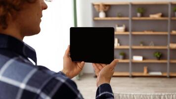 Man Holding Digital Tablet With Empty Screen Over Domestic Living Room Background, Standing At Home. Mockup For Smart House Application Or Website Advertisement Concept. Back View, Cropped, Panorama photo