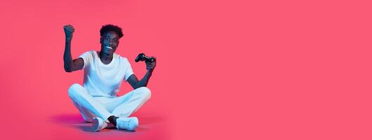 A young black man sits cross-legged on the floor, smiling and raising his fist in triumph while holding a gaming controller. The vibrant pink background adds energy to the joyful moment. photo