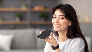 Digital Speaker Application. Portrait of young smiling woman sitting at table, talking on mobile phone, using laptop and virtual voice assistant for online search, to record reminder, dictate message photo