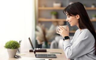 People And Technology Concept. Profile side view of smiling young woman using laptop and holding cup and drinking coffee. Female sitting at table, watching , movie or webinar, taking break photo