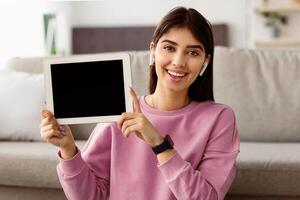 Great Application. Portrait of cheerful young lady in earphones holding and showing tablet with black empty screen for mockup, presenting gadget screen with blank space for template, copy space photo