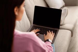 Over the shoulder shot of woman resting on couch in living room, using newest laptop with blank screen, websurfing or checking social media, chatting with friends, mockup, copy space photo