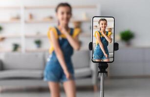 Young Blogger Concept. Cheerful teen girl dancing at camera filming using phone on tripod at home, creating her trendy content on a mobile app to share on social media, selective focus on screen photo