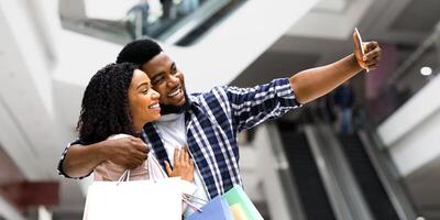 Positive Black Spouses Shopping In Department Store And Taking Selfie On Smartphone, African Couple Having Fun In Mall, Enjoying Doing Purchases Together, Carrying Colorful Shopper Bags, Free Space photo