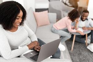 Smiling Black Woman Working Online On Laptop, Sitting On Couch With Computer Reading Email, Enjoying Freelance Work. Young Father Sitting On Floor Spending Time With Daughter Drawing On Table photo