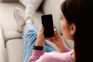 Over The Shoulder View Of Unrecognizable Young Woman Holding And Using Cell Phone, Sitting And Resting On The Couch In Living Room At Home. Selective Focus On Gadget, Template photo