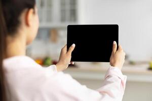 Over The Shoulder View Of Unrecognizable Woman Using Digital Tablet With Empty Screen Standing In Kitchen At Home. Application Or Internet Website Advertising, Mock Up, Free Space, Selective Focus photo