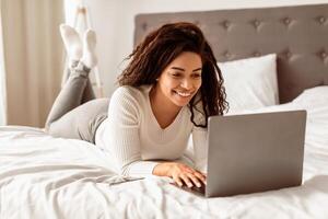 Portrait of happy black woman lying in bed and smiling, using laptop computer, chatting or browsing social media. Cheerful female freelancer working remotely at home office, typing on keyboard photo