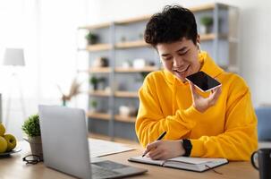 Work From Home. Young Asian Guy Talking On Mobile Phone And Taking Notes, Using Voice Assistant, Writing In Notebook. Smiling Man Sitting At Desk With Laptop At Home Office, Wearing Earbuds photo