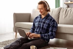 Smiling Man In Headphones Using Laptop Making Call Or Working Online Sitting On Floor At Home. Remote Job And Modern Communication, Internet And Technology Concept photo