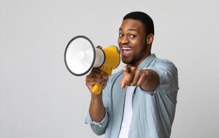 Positive african american guy shouting in megaphone and pointing at camera over grey background, copy space. Happy black man screaming with loudspeaker, cheering up, making advertisement photo