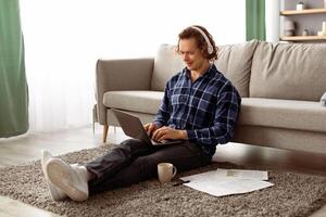 Happy Man Working On Laptop Wearing Headphones Browsing Internet Sitting On Floor At Home. Freelancer Guy Using Computer In Living Room. Distant Job, Internet And Technology photo