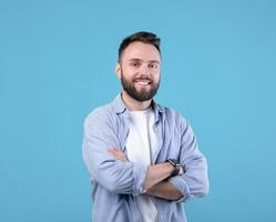 Portrait of positive young guy posing with crossed arms and smiling at camera on blue studio background. Masculine bearded millennial man in casual clothes having happy face expression photo