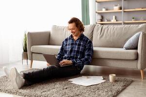 Distant Work. Happy Freelancer Man Using Laptop Working Online Sitting On Floor At Home. Relaxed Guy Browsing Internet On Computer In Living Room Indoor. Freelance, Remote Job, Internet And Technology photo