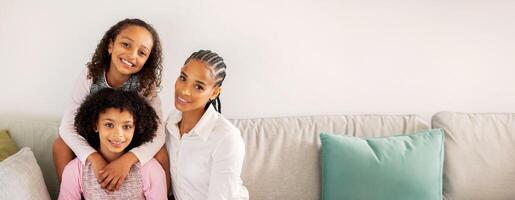 A black mother and her two daughters share a joyful moment in a cozy living room. They are smiling and posing closely together on a soft couch, surrounded by comfortable cushions. photo