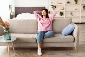Rest Concept. Beautiful lady sitting on comfortable couch at home in living room, looking aside at window. Happy woman relaxing on sofa, leaning back, enjoying weekend free time or break from work photo