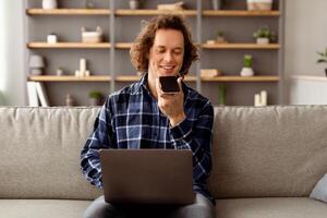 Man Using Voice Assistant On Smartphone Working On Laptop Sitting On Couch At Home. Modern Hipster Guy Talking With Loudspeaker Mode Holding Phone Near Mouth. Voice Search Mobile Application photo
