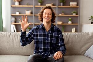 Hello Greeting. Cheerful Guy Waving Hand To Camera Sitting On Sofa At Home. Young Man Smiling And Gesturing Hi Posing Indoors. Call, Modern Distance Communication Concept photo