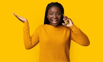Unexpected Call. Portrait Of Surprised Young Black Woman Talking On Cellphone And Emotionally Gesturing, African Lady Having Phone Conversation, Standing Over Yellow Background In Studio, Copy Space photo