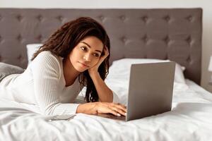 Portrait of African American woman lying in bed and using laptop computer, chatting or browsing social media. Young lady watching or movie, working remotely at home office, typing on keyboard photo