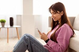 Scrolling News Feed. Side view profile portrait of young smiling lady holding mobile phone, typing sms message, sitting on floor and leaning on couch. Woman browsing internet, surfing web, using app photo