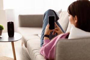 People And Technology Concept. Over The Shoulder View Of Unrecognizable Lady Using Mobile Phone With Black Empty Screen For Mockup, Holding Device, Lying On Sofa, Blurred Background, Selective Focus photo